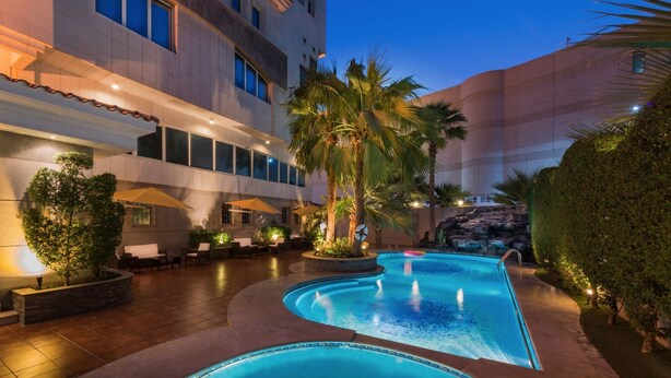 Zona de piscina de hotel con piscina curva, palmeras y mobiliario exterior.