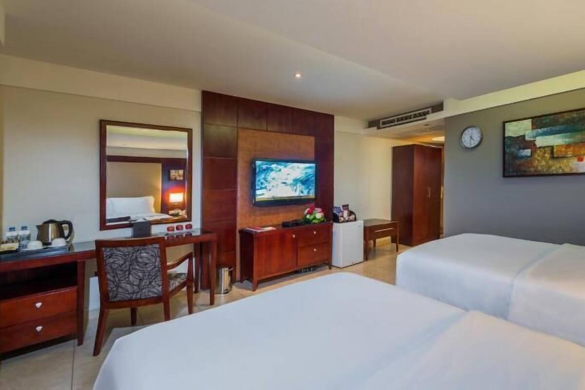 A hotel room with two beds, a desk with a chair, a television, a clock, and a painting on the wall.