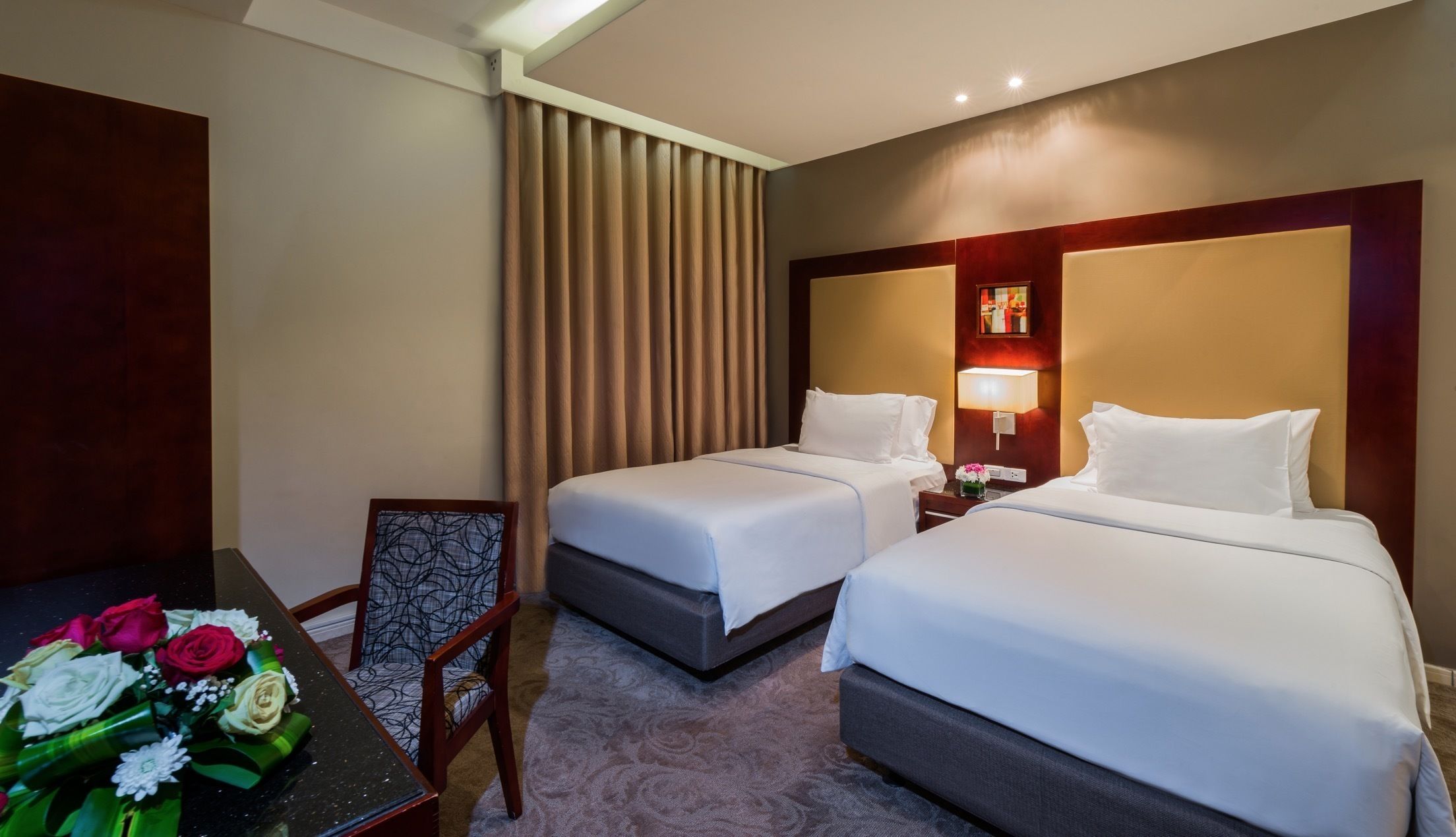 A hotel room with two beds, a desk with a chair, a vase of flowers, and a large window with curtains.
