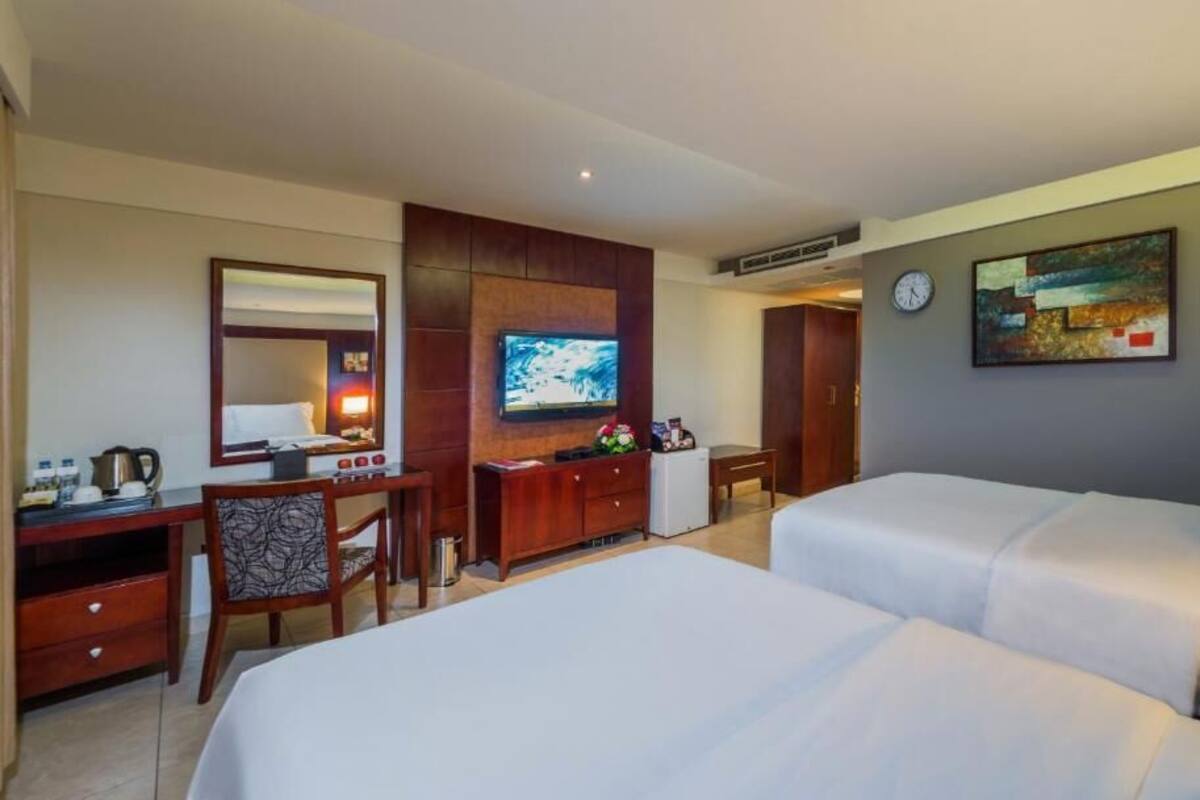A hotel room with two beds, a desk with a computer, a TV, a clock, and a painting on the wall.