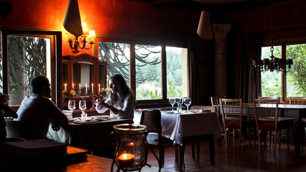 A dimly lit restaurant with wooden interiors, large windows offering a view of greenery, and candlelit tables. - Los Juncos Patagonian Lake House (San Carlos de Bariloche)