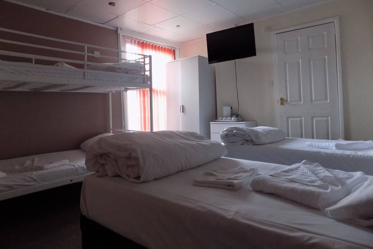 Una habitación de hotel con literas, un televisor de pantalla plana montado en la pared, una mesita de noche con lámpara y una puerta.