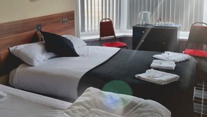A hotel room with two beds, a desk, and a chair. There is a window with blinds, a painting on the wall, and a view of the outdoors. - The New Royal Windsor Hotel (Blackpool)