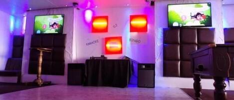 Een kamer met een podium, karaoke-installatie en grote schermen waarop een cartoonfiguur wordt getoond.