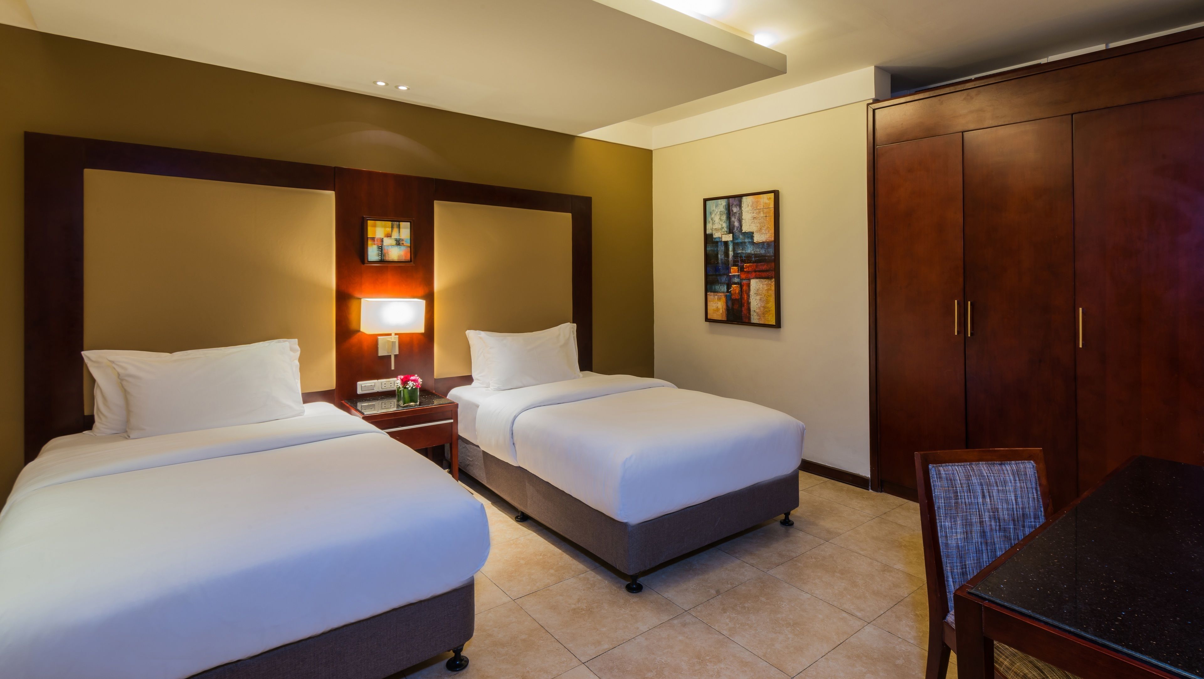 A hotel room with two beds, a nightstand with a lamp, a painting on the wall, and a wardrobe.