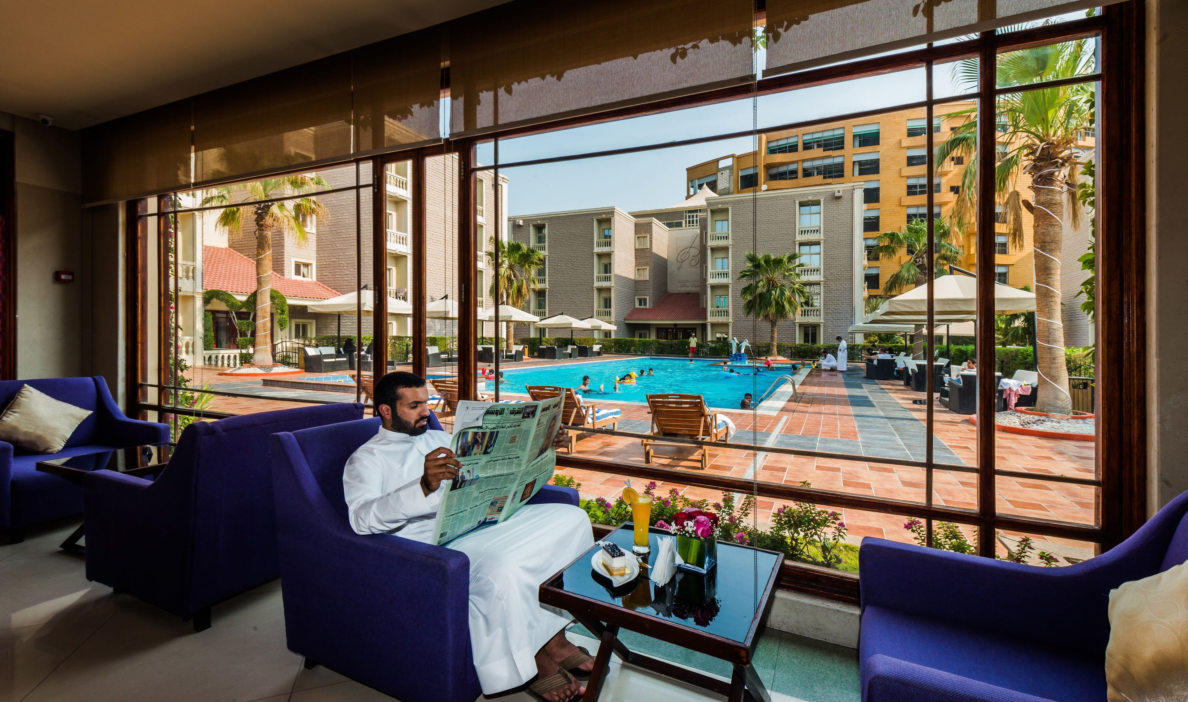 A hotel lobby with large windows overlooking a swimming pool and a courtyard with palm trees and outdoor seating.