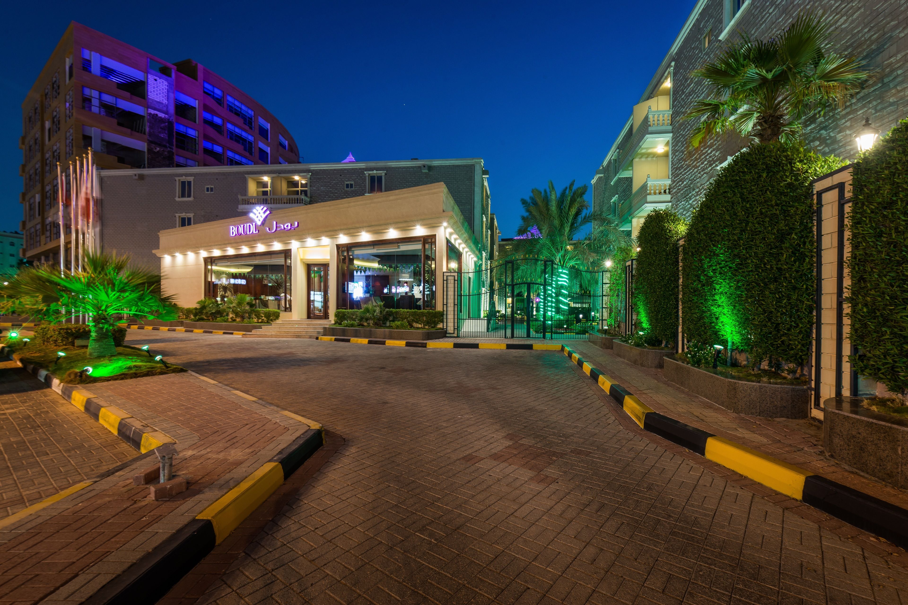 A hotel entrance with a modern design, featuring a well-lit facade and a landscaped area with palm trees and shrubs.