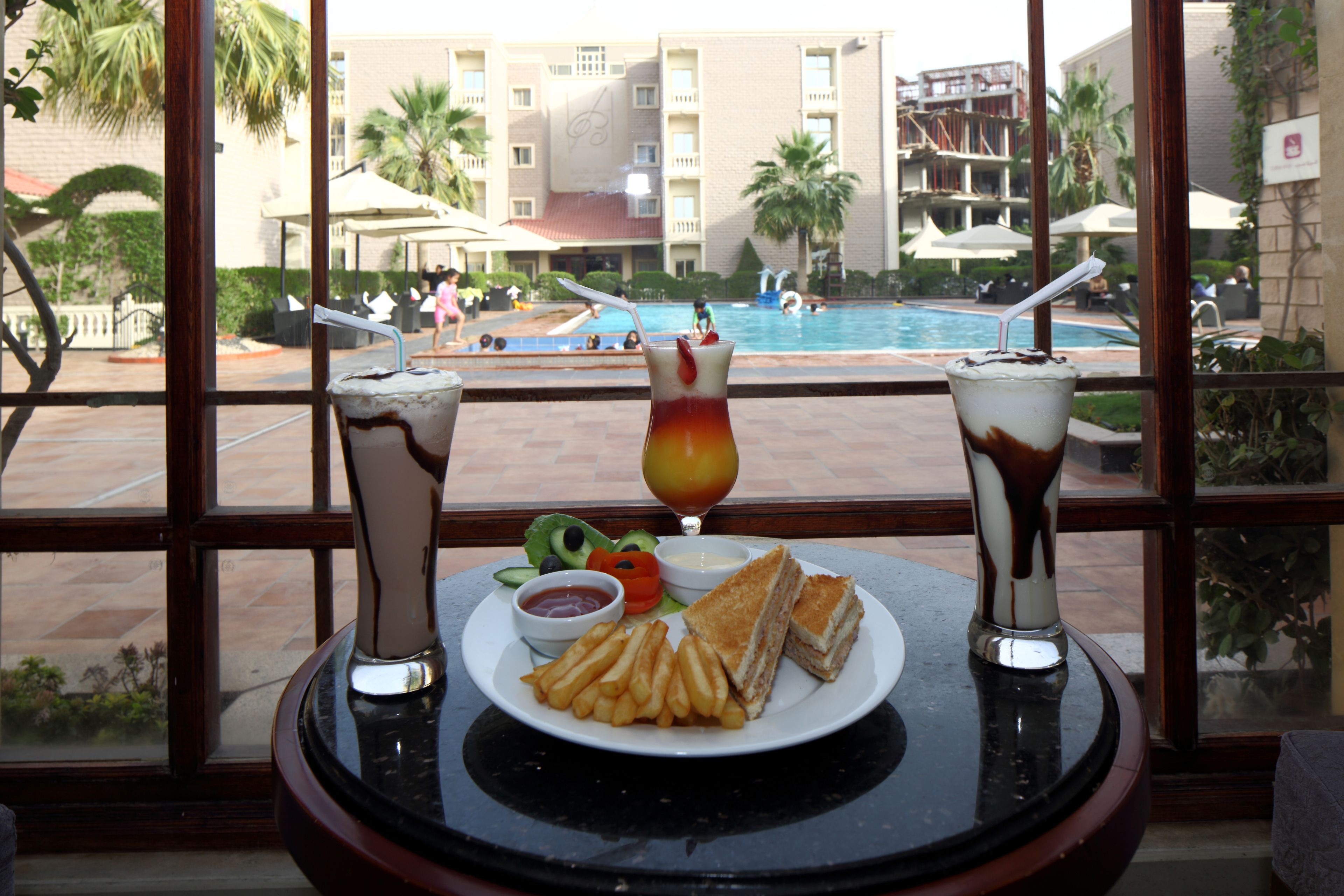 Una mesa con un plato de comida, que incluye un sándwich, papas fritas y una bebida, con vista a una piscina al fondo.