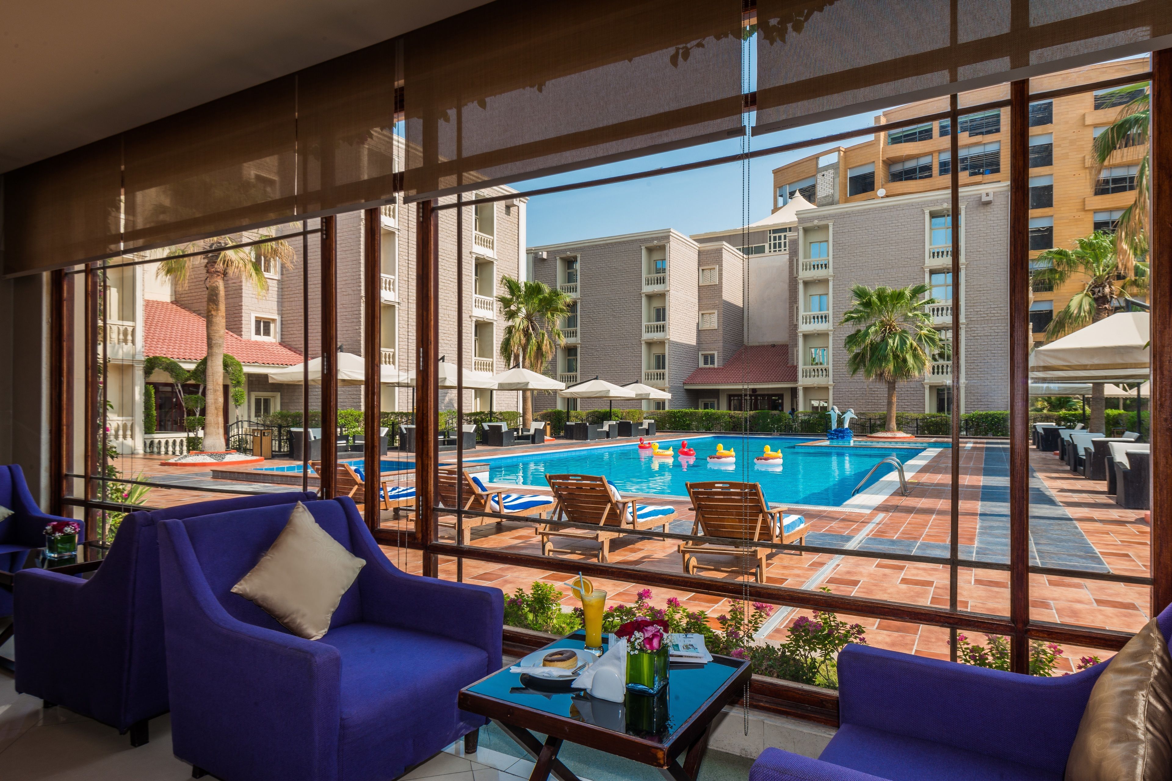 A poolside area with lounge chairs, a table with a drink, and a view of a building with balconies and palm trees.