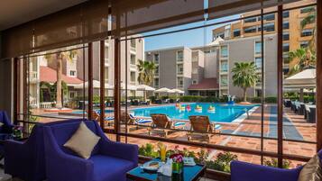 A poolside area with lounge chairs, a table with a drink, and a view of a building with balconies and palm trees.