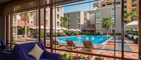 A poolside area with lounge chairs, a table with a drink, and a view of a building with balconies and palm trees.