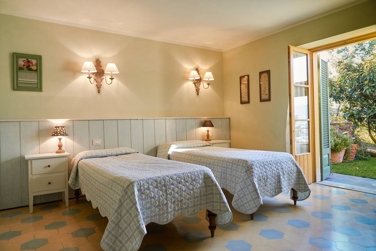 Een kamer met twee bedden, een nachtkastje met een lamp, wandlampen en een deur naar een tuin.