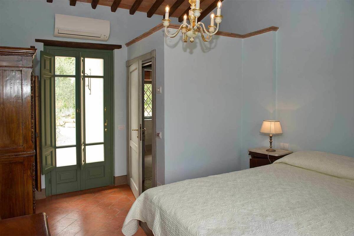 Een slaapkamer met een bed, nachtkastje, een kroonluchter, een houten kledingkast en een deur naar buiten.