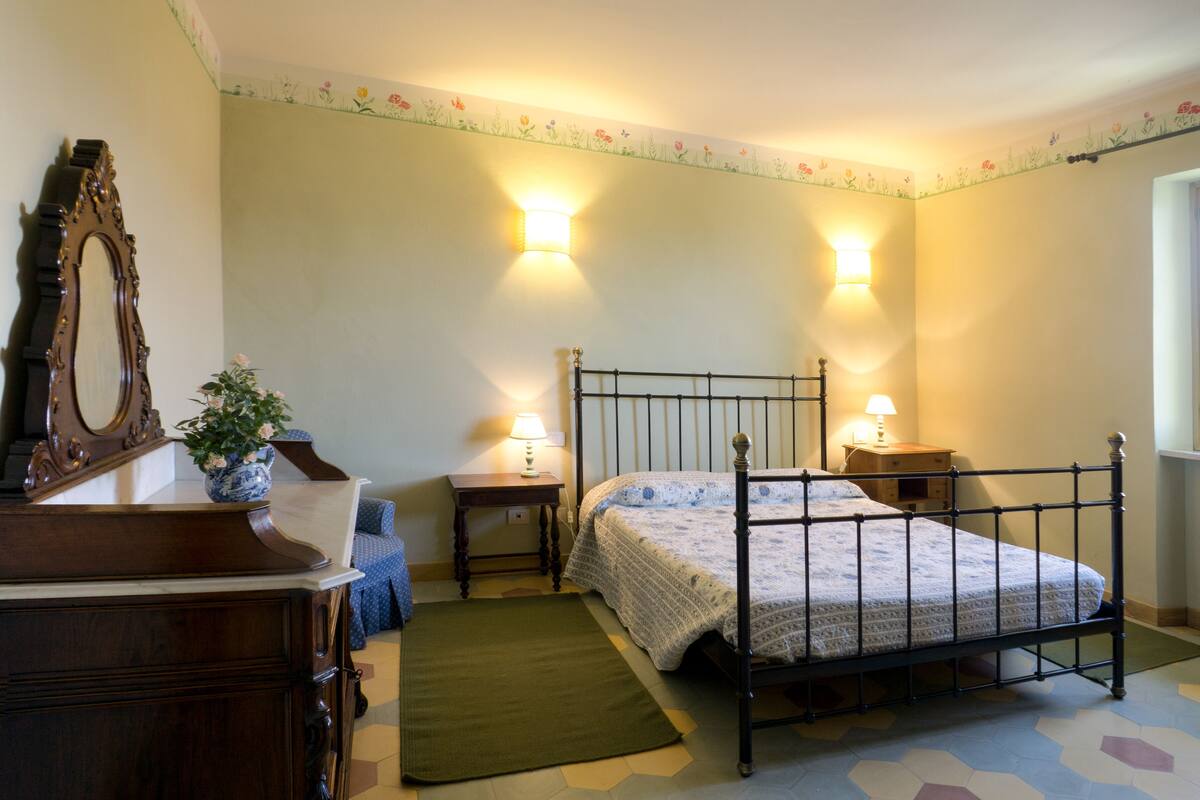Een slaapkamer met een hemelbed, twee nachtkastjes met lampen, een kaptafel, een spiegel en een bloemenbehang.
