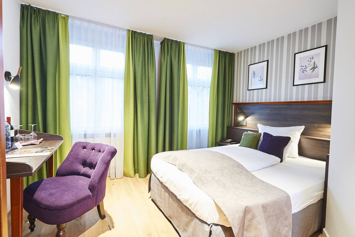 Quarto de hotel com cama, poltrona, escrivaninha e cortinas.