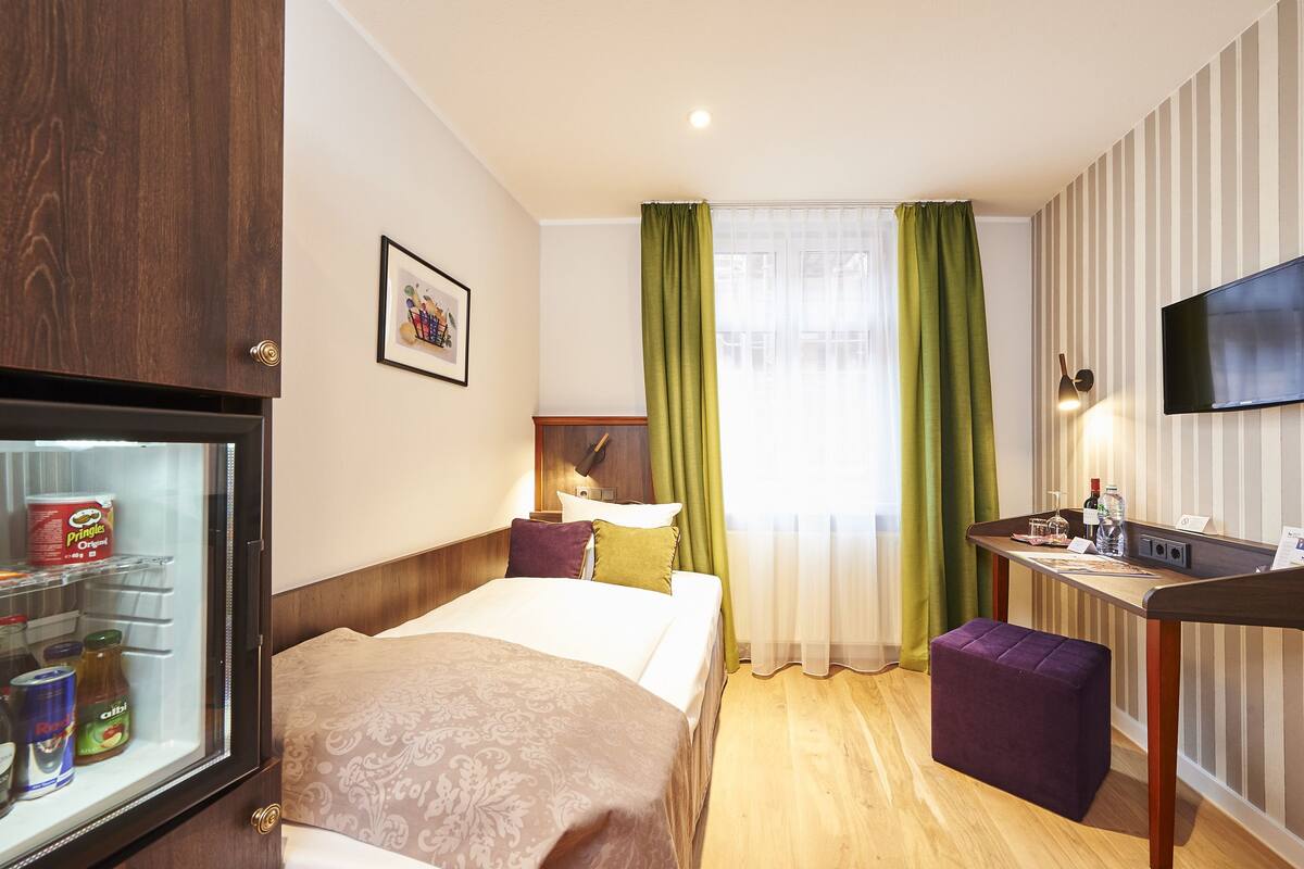 Quarto de hotel com cama, secretária, televisão, minibar e janela com cortinas.