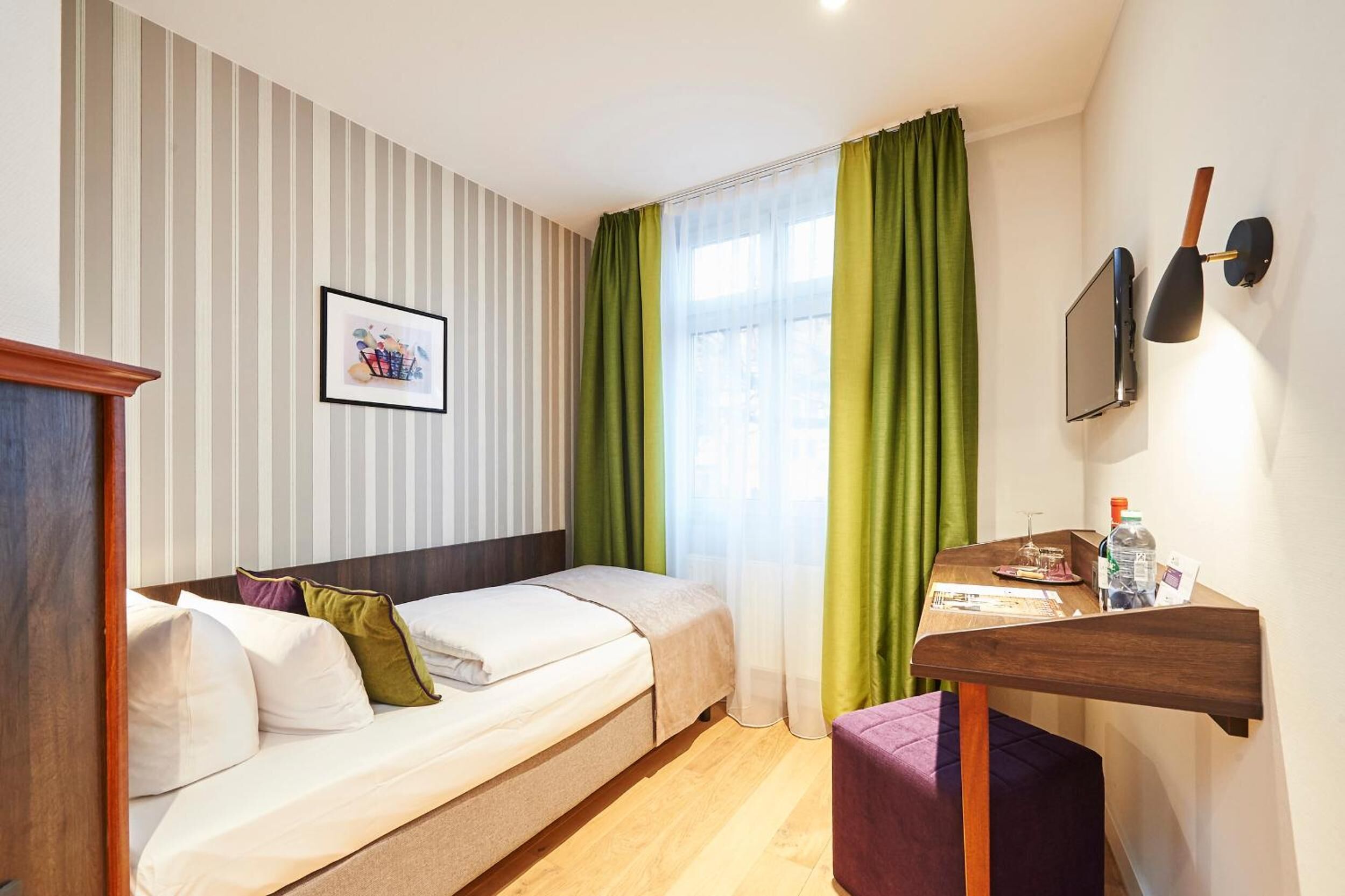 A compact hotel room with a single bed, a small desk with a chair, a mounted TV, and a window with green curtains.