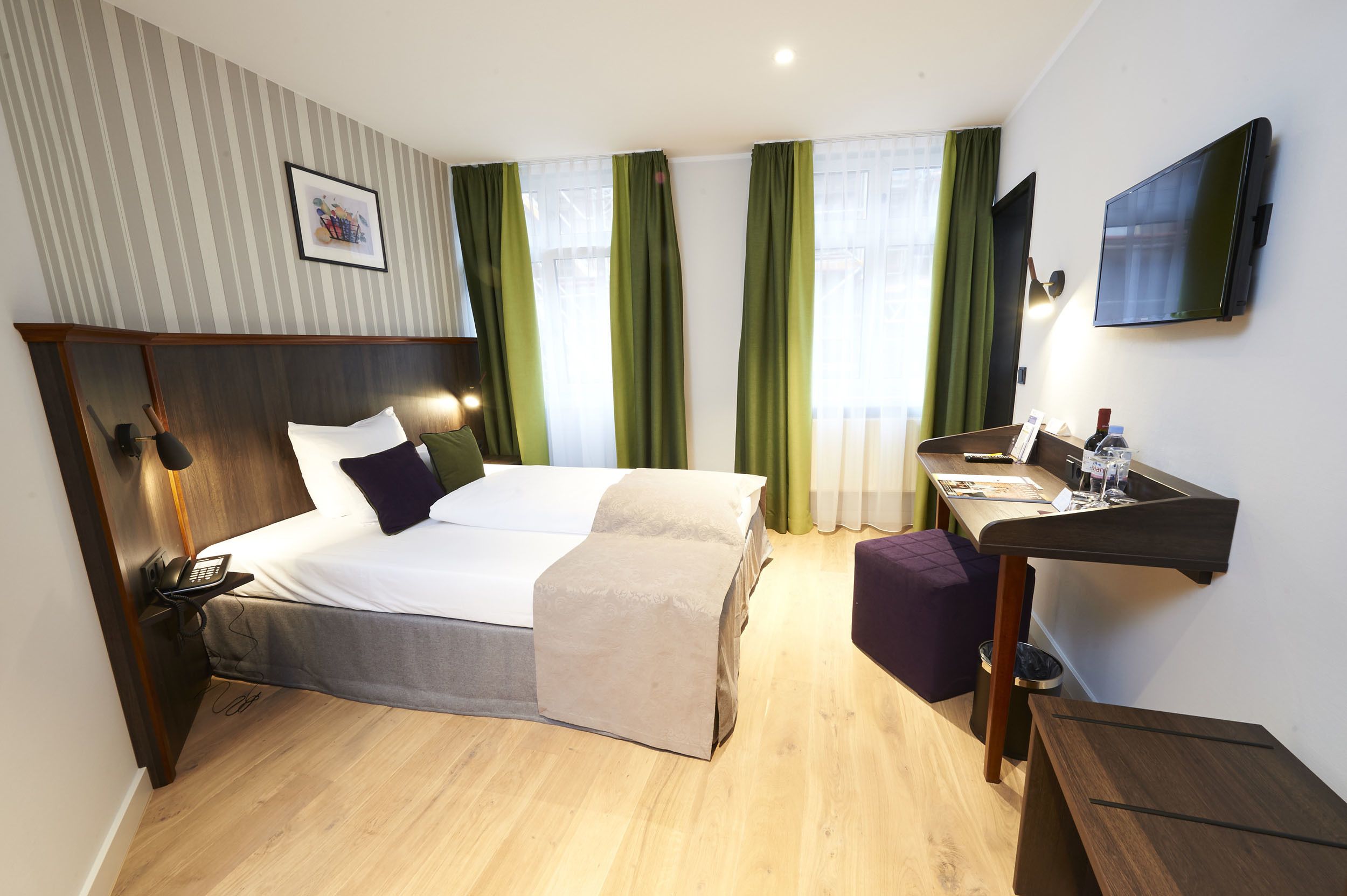 A hotel room with a bed, bedside table with a phone, a desk with a chair, a TV, and a window with curtains.