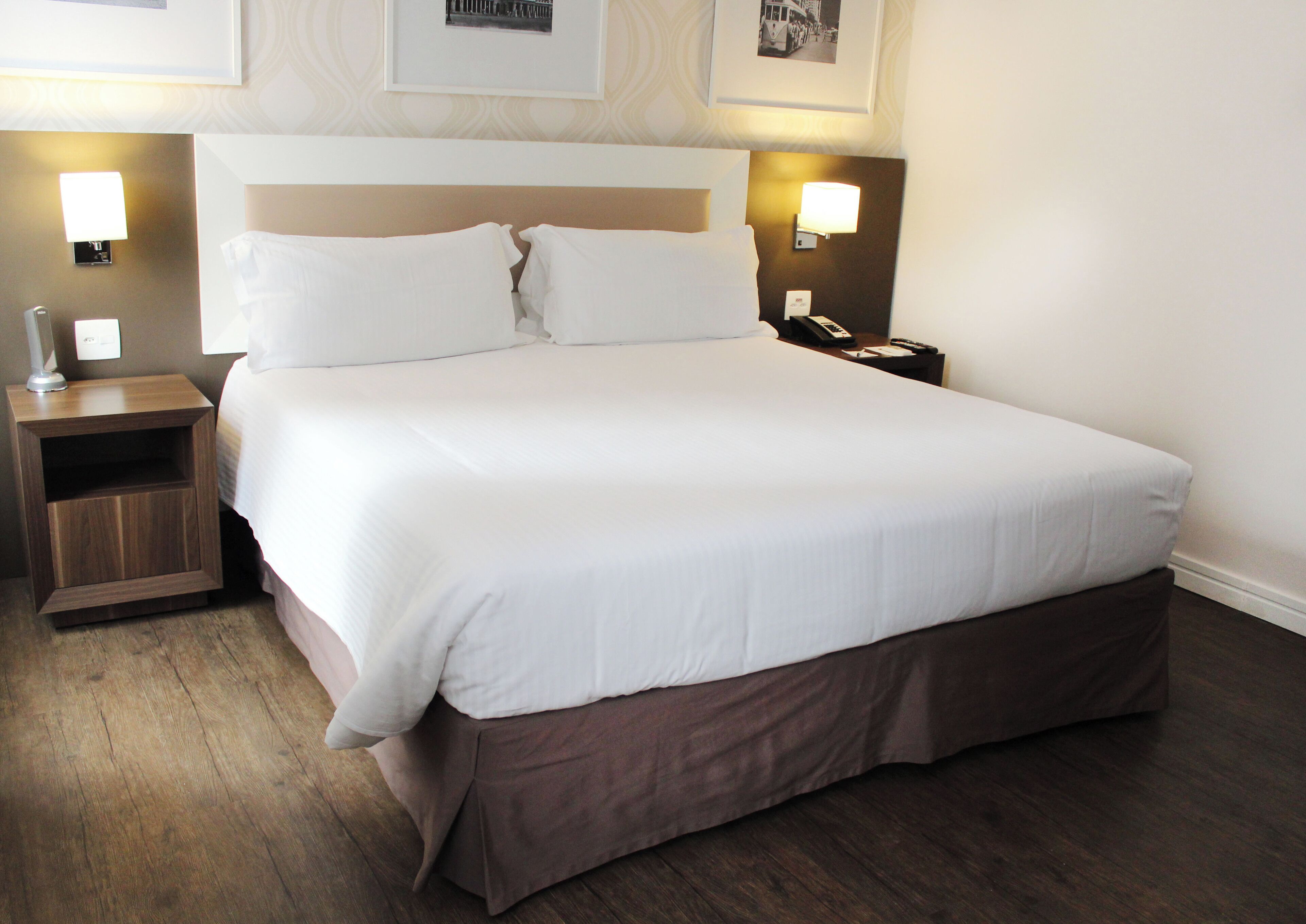 A neatly made bed with white linens, a wooden nightstand with a lamp, and a bedside table with a phone and remote control.