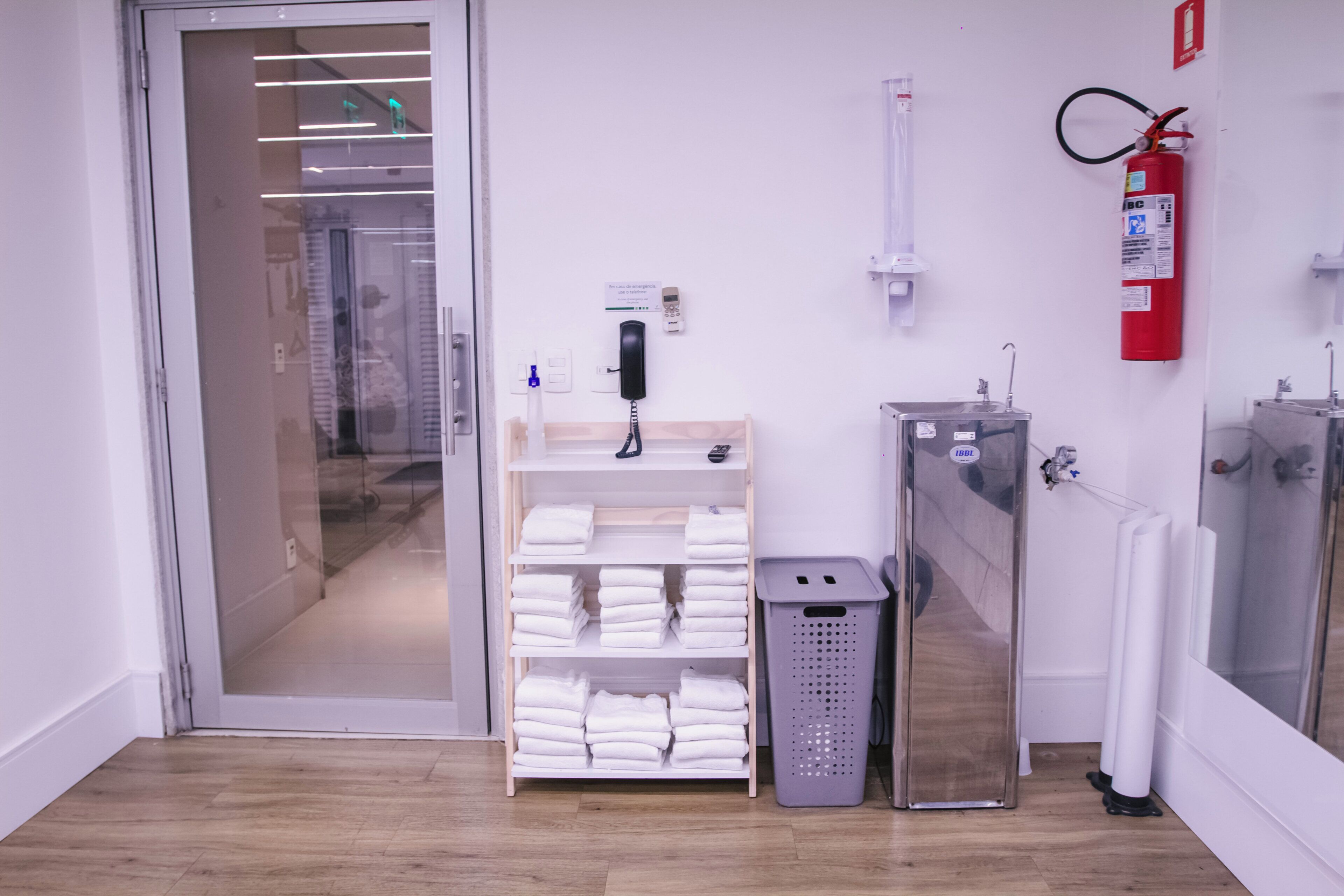 A hotel room with a glass door leading to a hallway, a white shelf with folded towels, a trash bin, a fire extinguisher, and a radiator.
