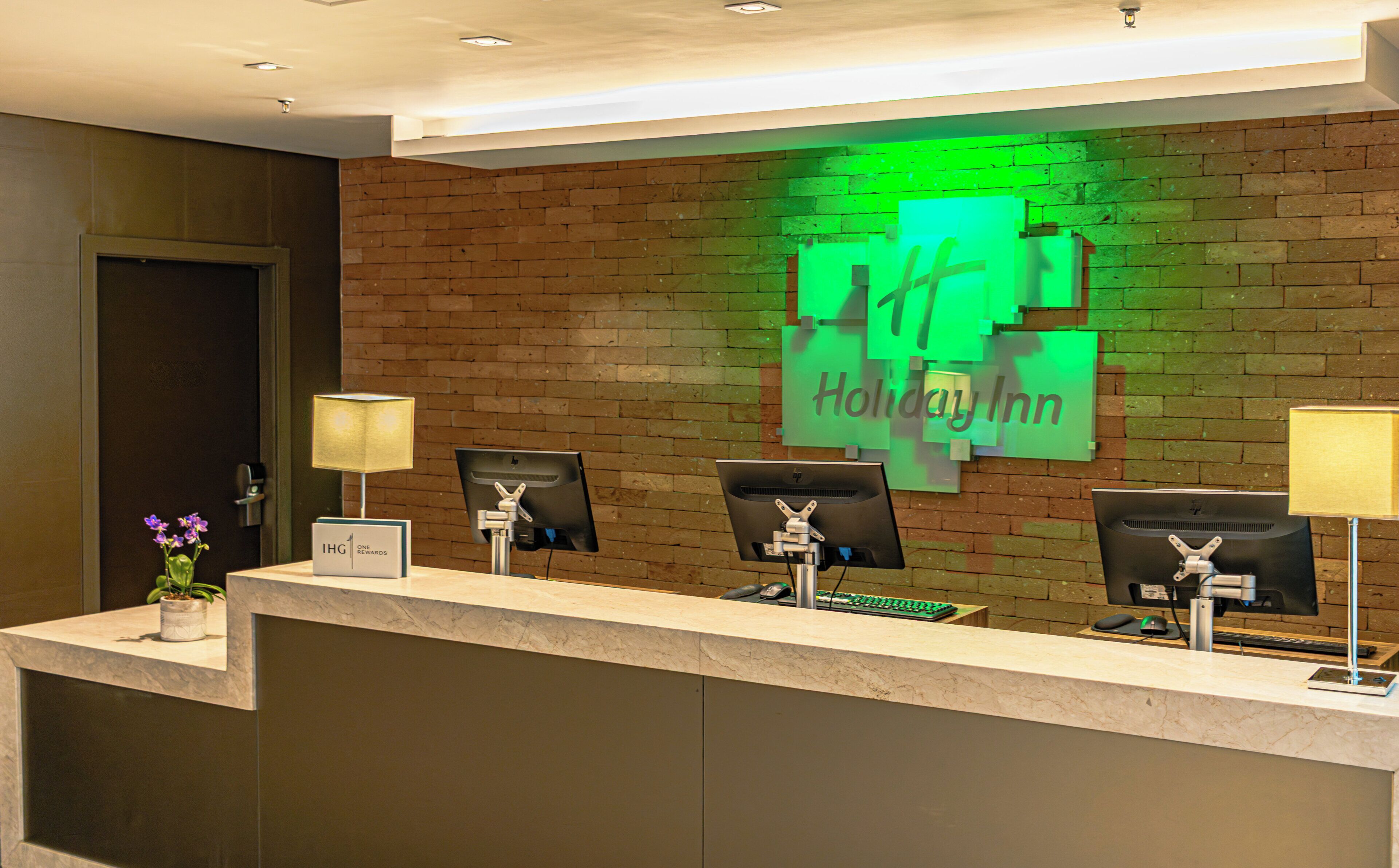 Una reception hotel con un muro in mattoni, tre monitor per computer e un cartello con la scritta Holiday Inn.