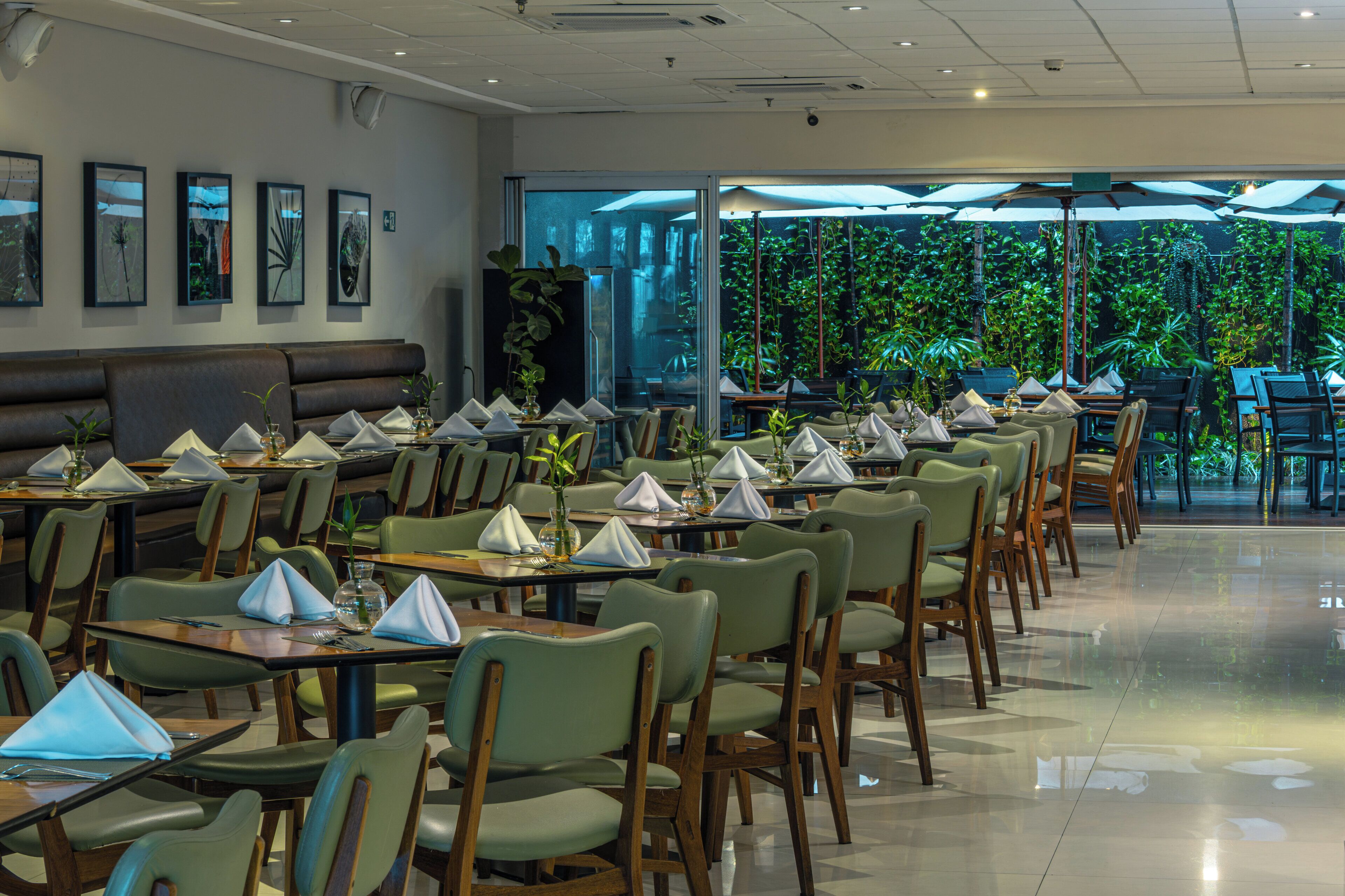 Restaurante com mesas preparadas para refeições, cadeiras e uma grande janela com vista para o verde.