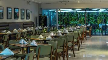 Restaurante com mesas preparadas para refeições, cadeiras e uma grande janela com vista para o verde.