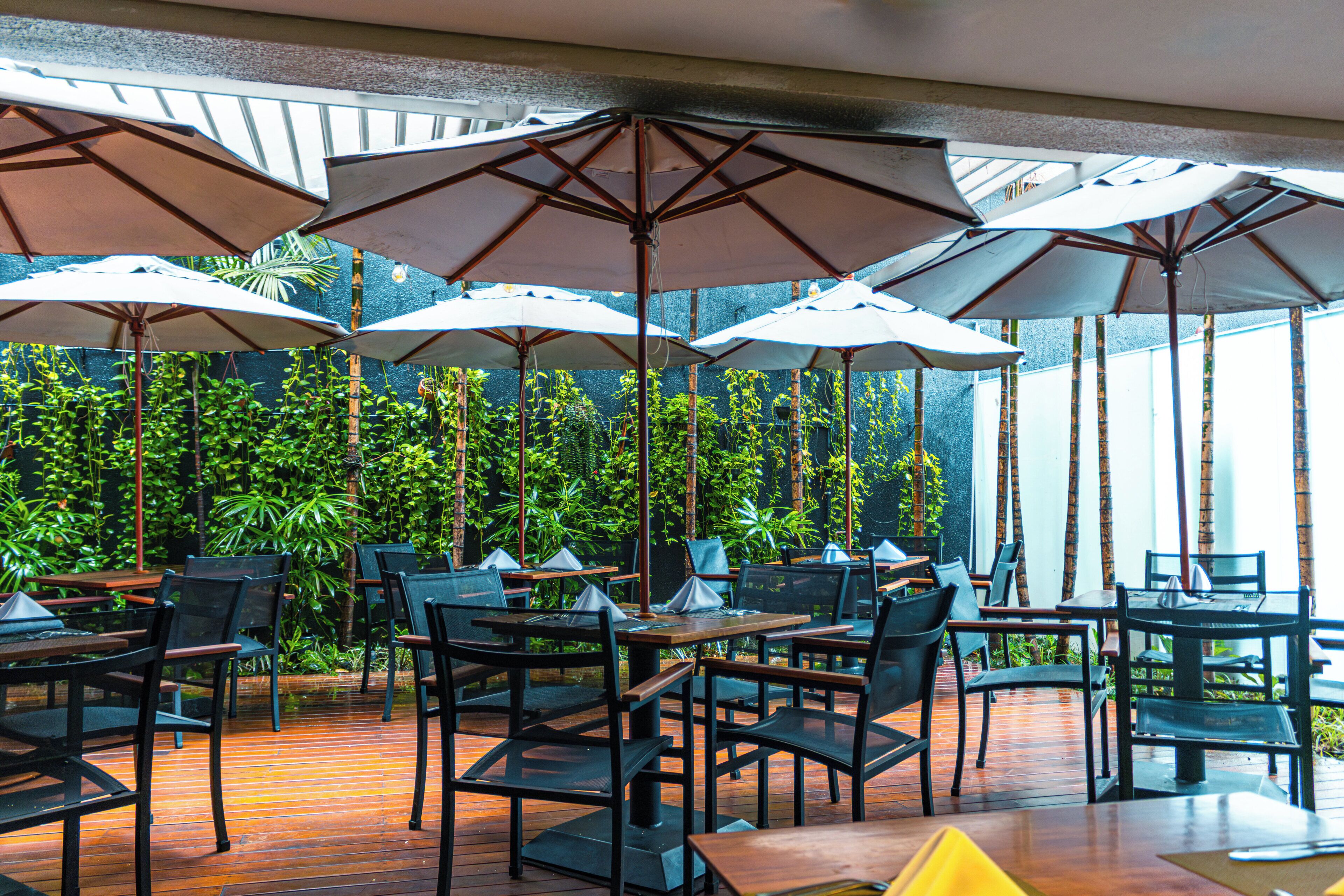 An outdoor dining area with multiple tables and chairs, surrounded by lush greenery and large umbrellas.