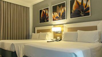 Quarto de hotel com duas camas, uma mesinha de cabeceira, um abajur e três quadros decorativos com flores na parede.