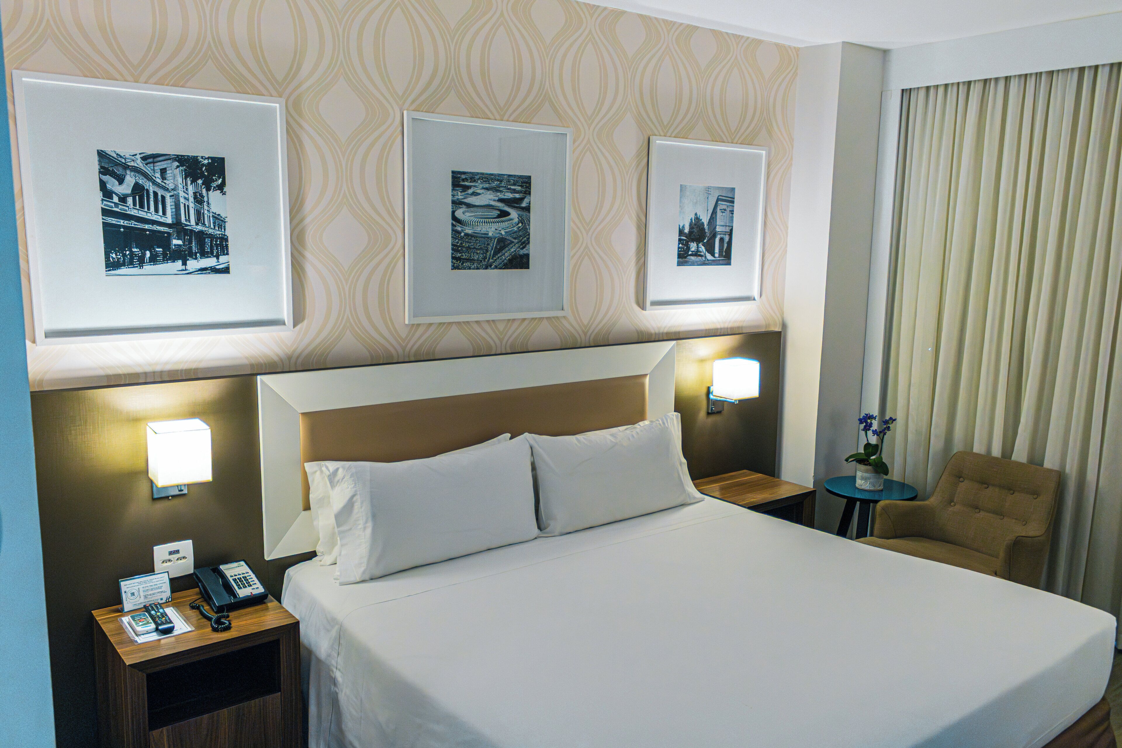 Habitación de hotel con una cama, dos mesitas de noche, una silla y tres cuadros en la pared.