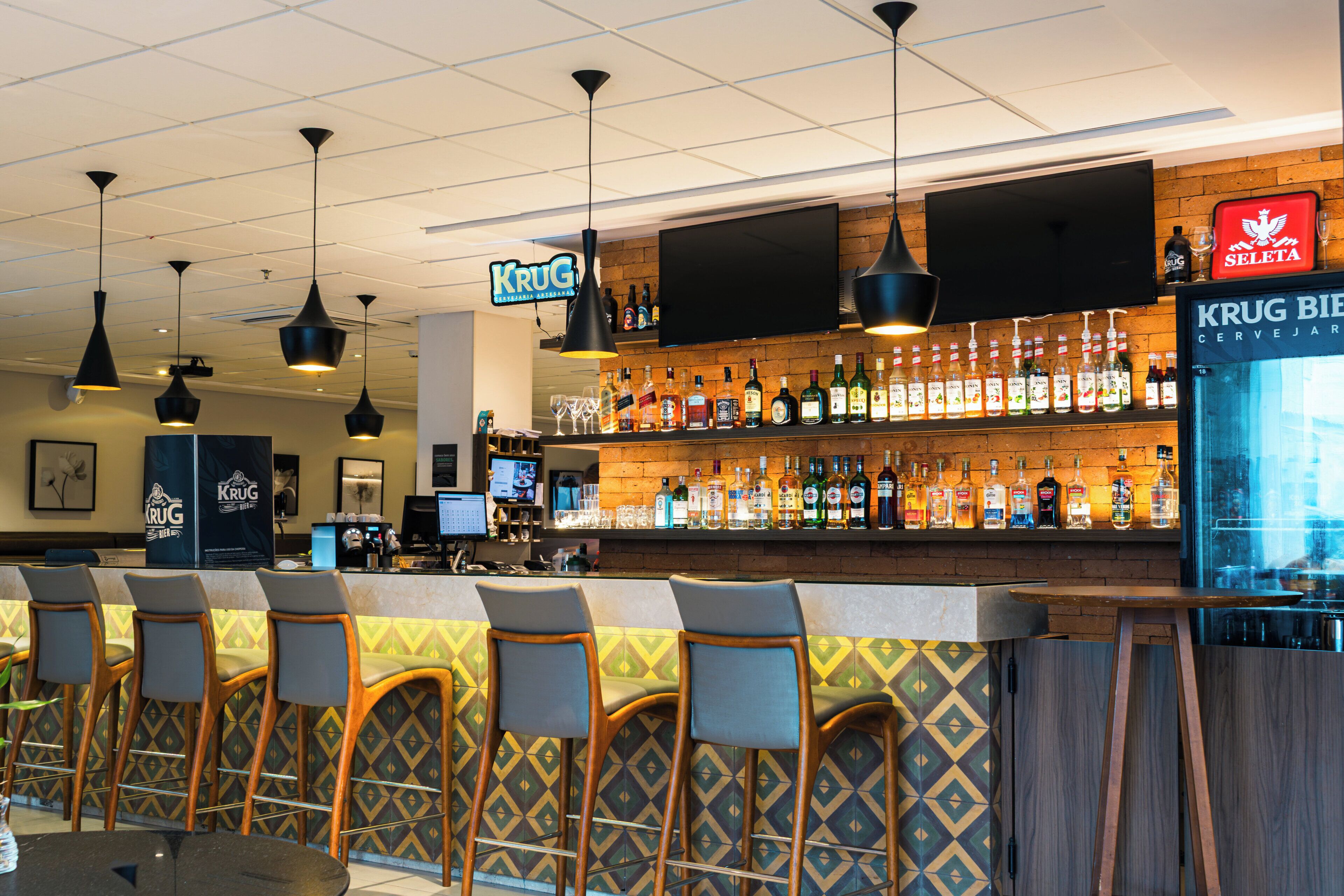 Zona de bar con mostrador, taburetes, televisor montado en la pared y una variedad de botellas y vasos en estanterías.