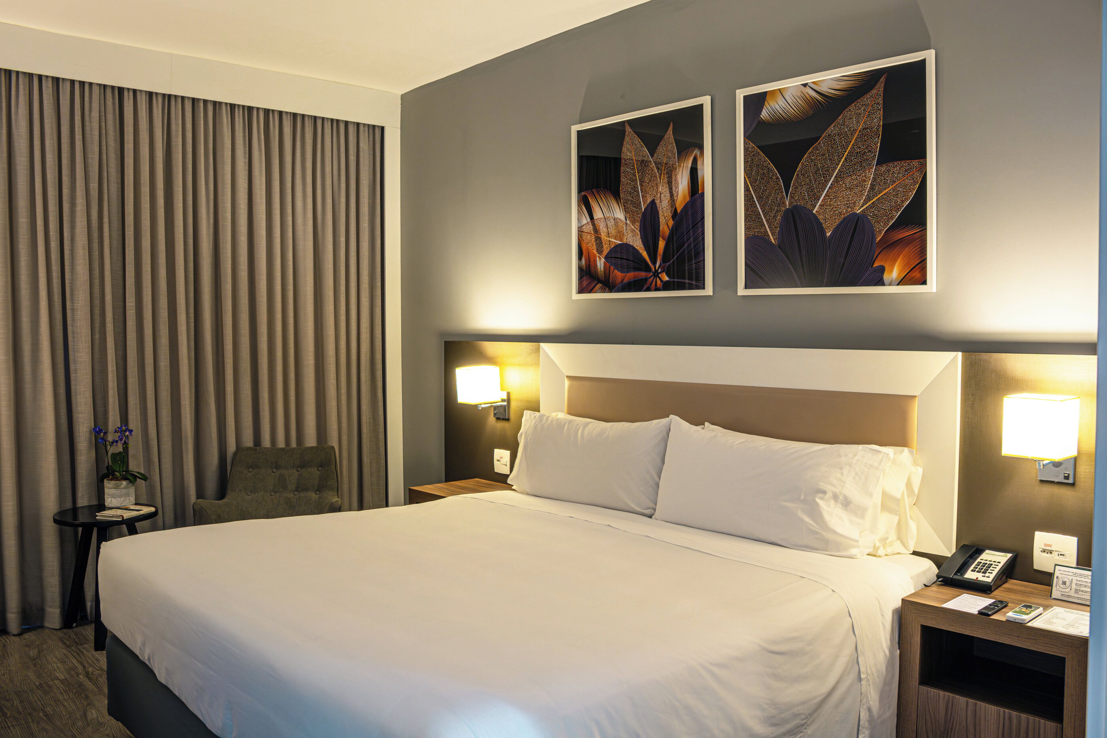 Quarto de hotel com uma cama grande, uma cadeira, mesa de cabeceira com telefone e abajur, e duas luminárias de parede.