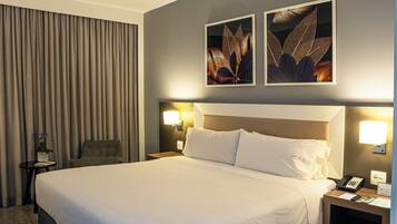 Quarto de hotel com uma cama grande, uma cadeira, mesa de cabeceira com telefone e abajur, e duas luminárias de parede.