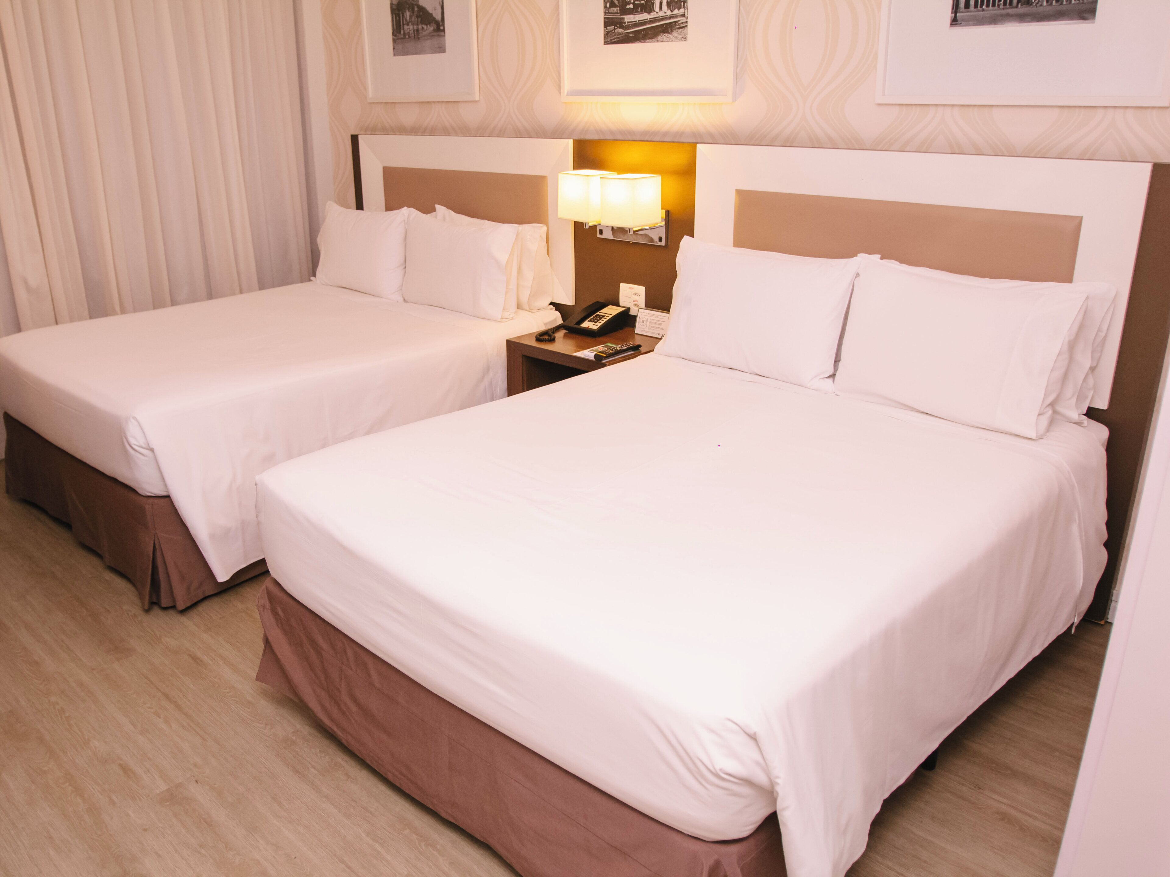 Quarto de hotel com duas camas, uma mesa de cabeceira com abajur e quadros nas paredes.
