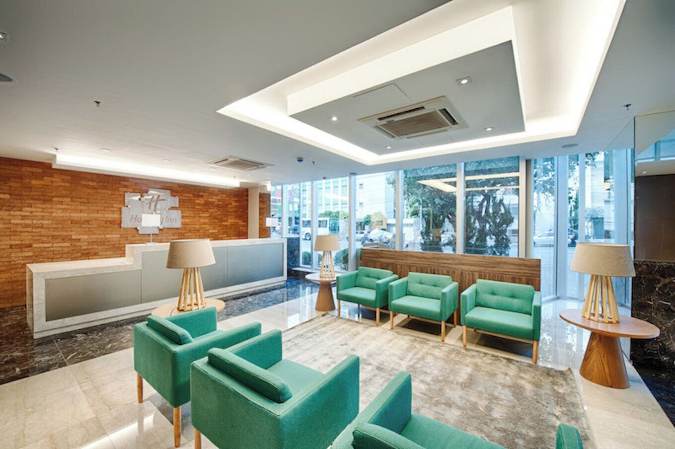 A modern lobby with a reception desk, green seating area, and large windows offering a view of the outdoors.