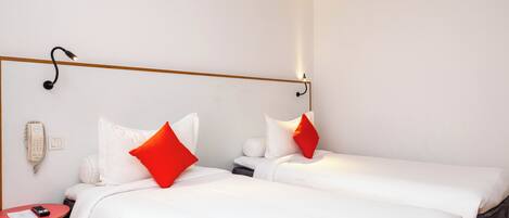 A hotel room with two beds, each with white bedding and red pillows, a bedside table with a remote control, and a wall-mounted phone.