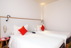 Camera d'albergo con due letti, entrambi con biancheria da letto bianca e cuscini rossi, un comodino con telecomando e un telefono fissato al muro.