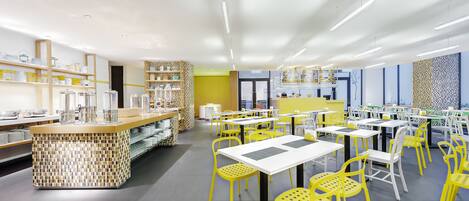 Un restaurante muy iluminado con sillas amarillas y mesas blancas, que presenta una isla central con base de mosaicos.
