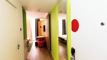 A hotel room with a wooden floor, a bed, a small table, and a red ottoman.