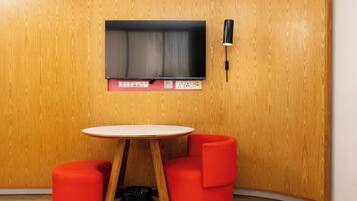 A room with a wooden wall, a flat-screen TV mounted above a table with two red chairs, and a black trash bin.