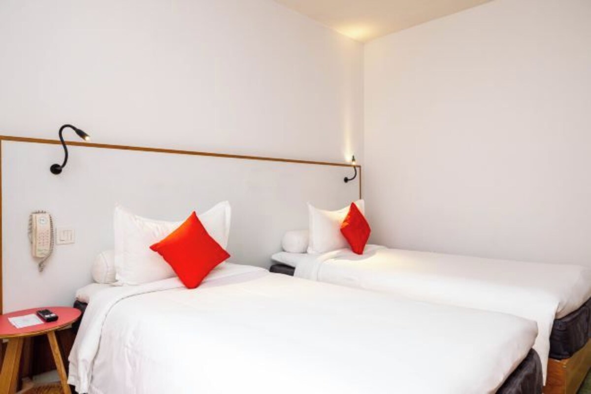 Ett hotellrum med två sängar, vita sängkläder, röda kuddar, sängbord, en telefon och en väggmonterad lampa.