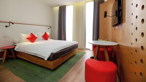 Una habitación de hotel moderna con una cama de madera, almohadas rojas y blancas, una alfombra verde, una pequeña mesa redonda y una silla roja.