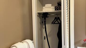 A compact bathroom with a towel rack, a vacuum cleaner, and a shelf with folded towels and a garment.