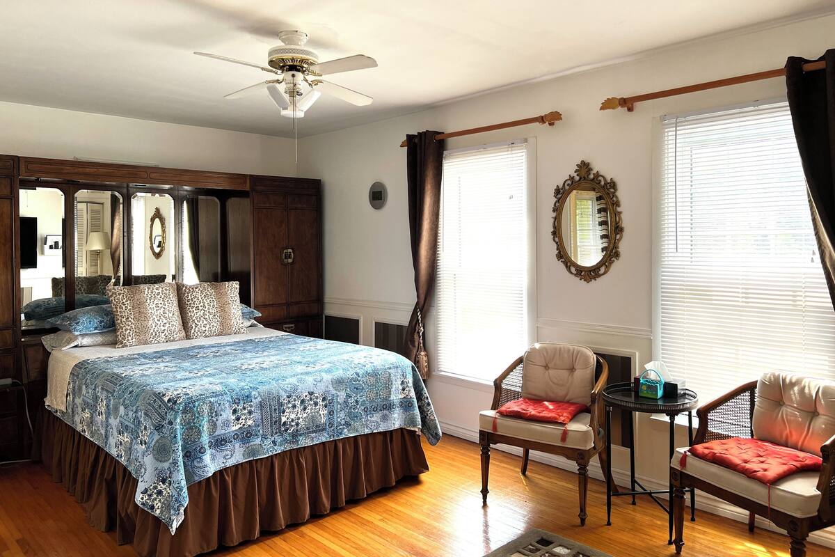 A bedroom with a bed, a chair, a table, a mirror, and a ceiling fan.
