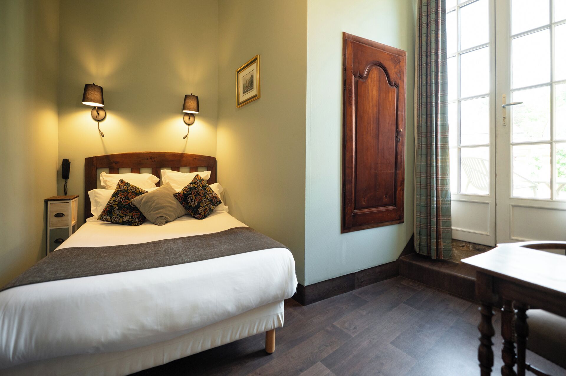 A neatly made bed with a wooden headboard, two bedside lamps, a bedside phone, and a window with curtains.