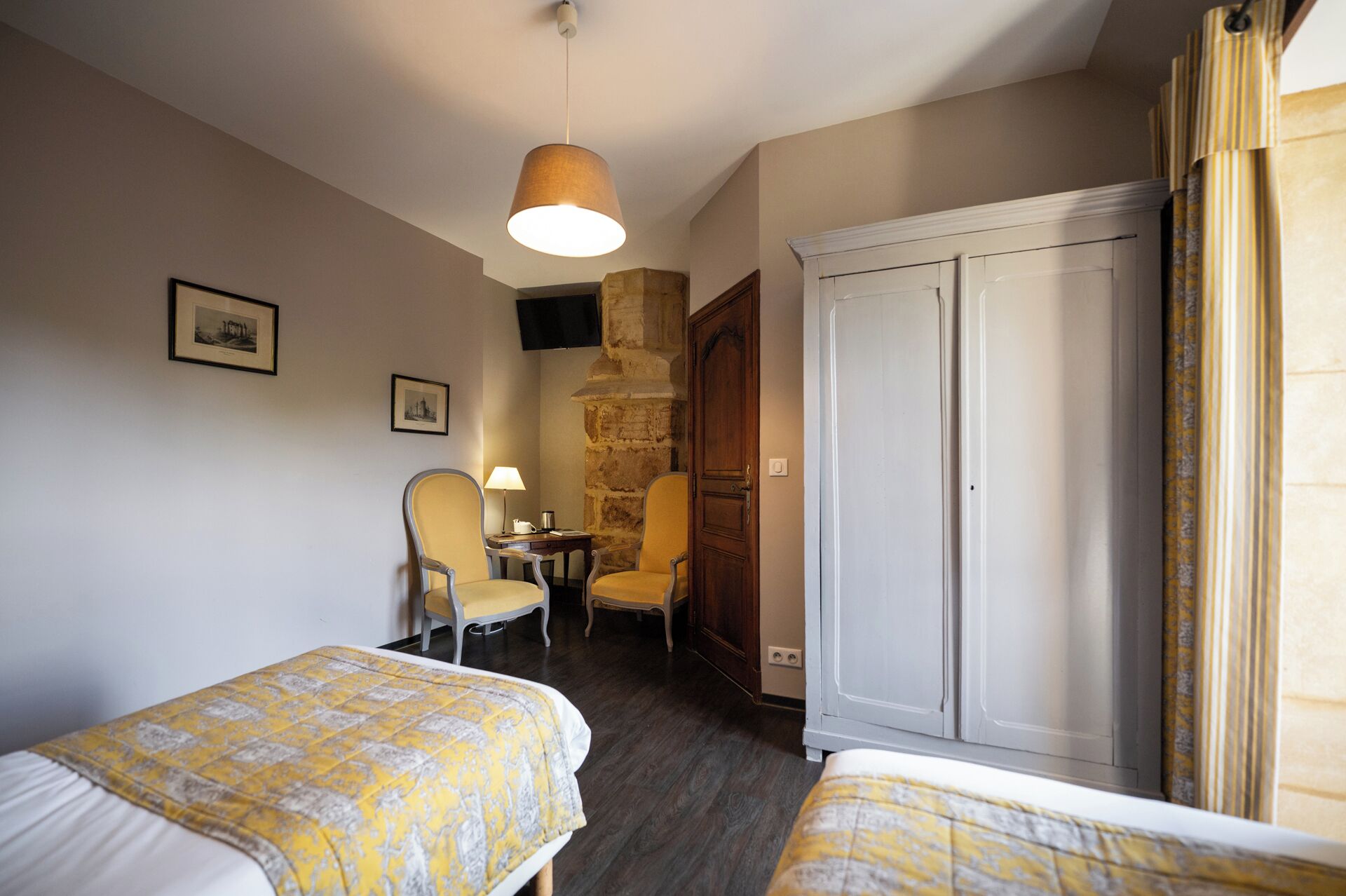 A hotel room with two beds, a stone wall, a wooden wardrobe, a chair, a table, and a hanging light fixture.