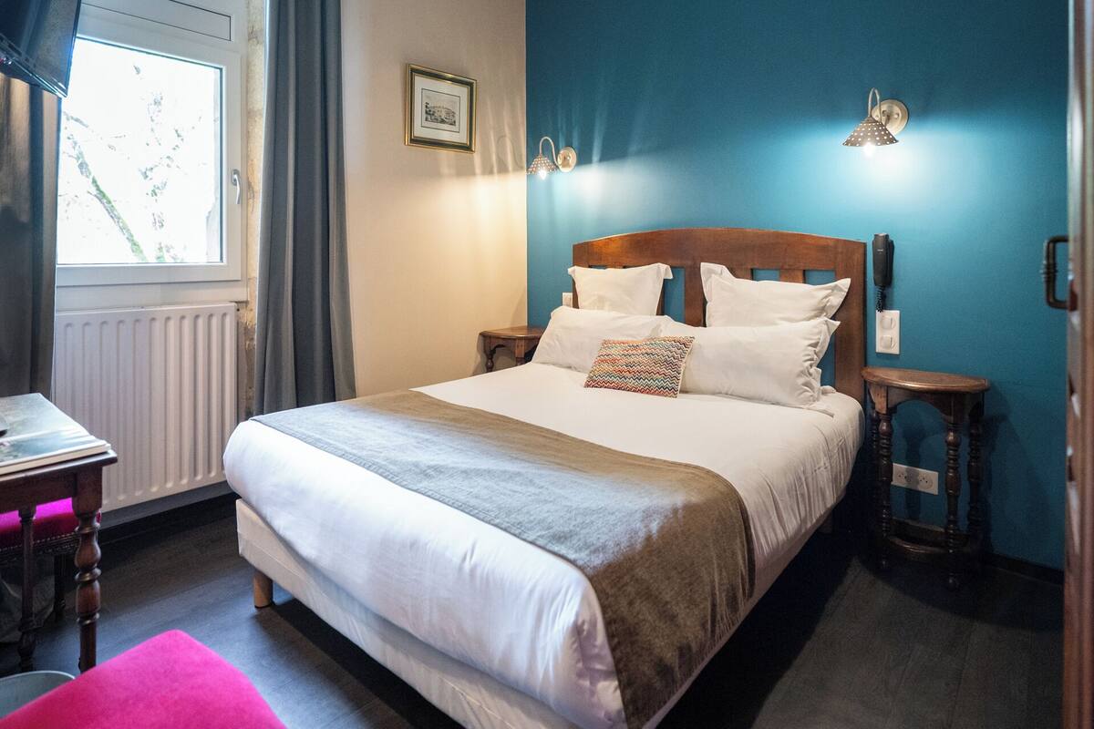 A neatly made bed with white linens and a patterned pillow, a wooden nightstand with a phone, a framed picture on the wall, and a window with curtains.