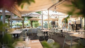 Een buitenruimte met eettafels en stoelen onder parasols, omgeven door groen en een stenen gebouw op de achtergrond.