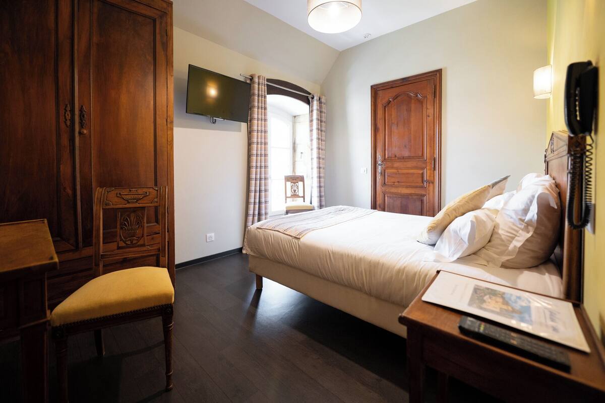 A bedroom with a bed, wooden wardrobe, a television, a chair, and a bedside table with a remote control.