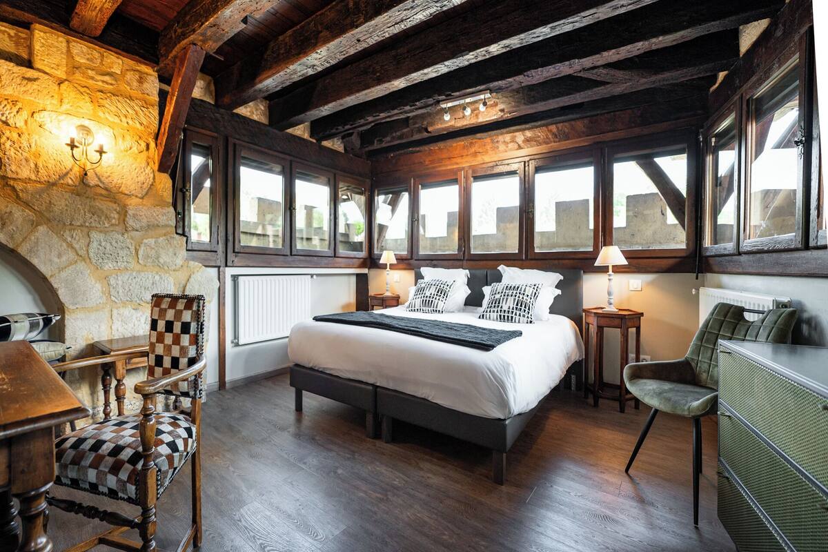 A bedroom with a large bed, wooden beams, stone walls, and a view through windows.